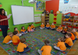 Dzieci w pomarańczowych koszulkach siedzą na dywanie, pani intendent przedstawia dzieciom właściwości odżywcze marchewki.